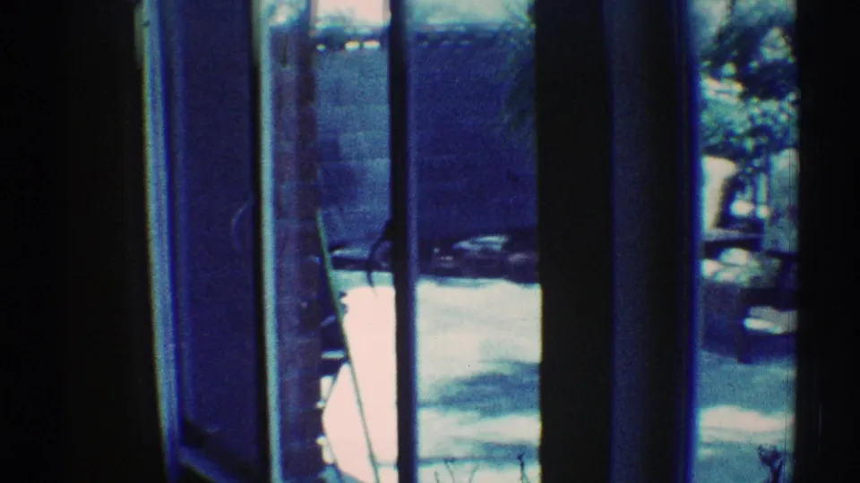 1969 Los Angeles 8mm frame looking through blue-tinted glass toward a patio chair, bright pavement, plants, and window framing.