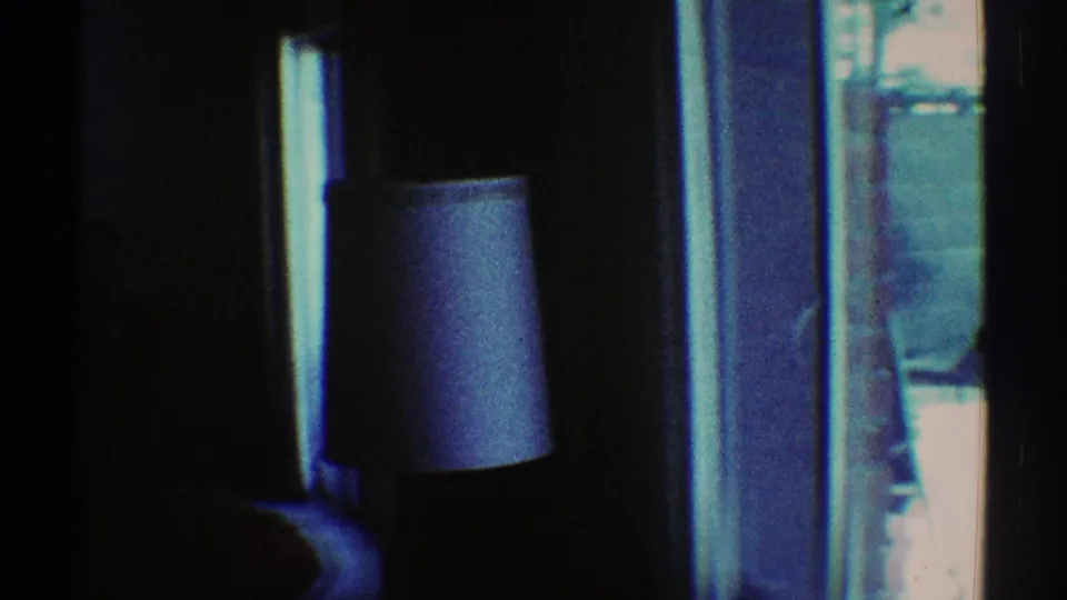 1969 Los Angeles 8mm frame showing a dim interior area with a lampshade near blue window glass and bright exterior light.