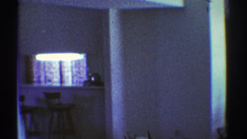 1969 apartment kitchen area with a bright pass-through counter, bar stools, and dark foreground walls in grainy 8mm film.