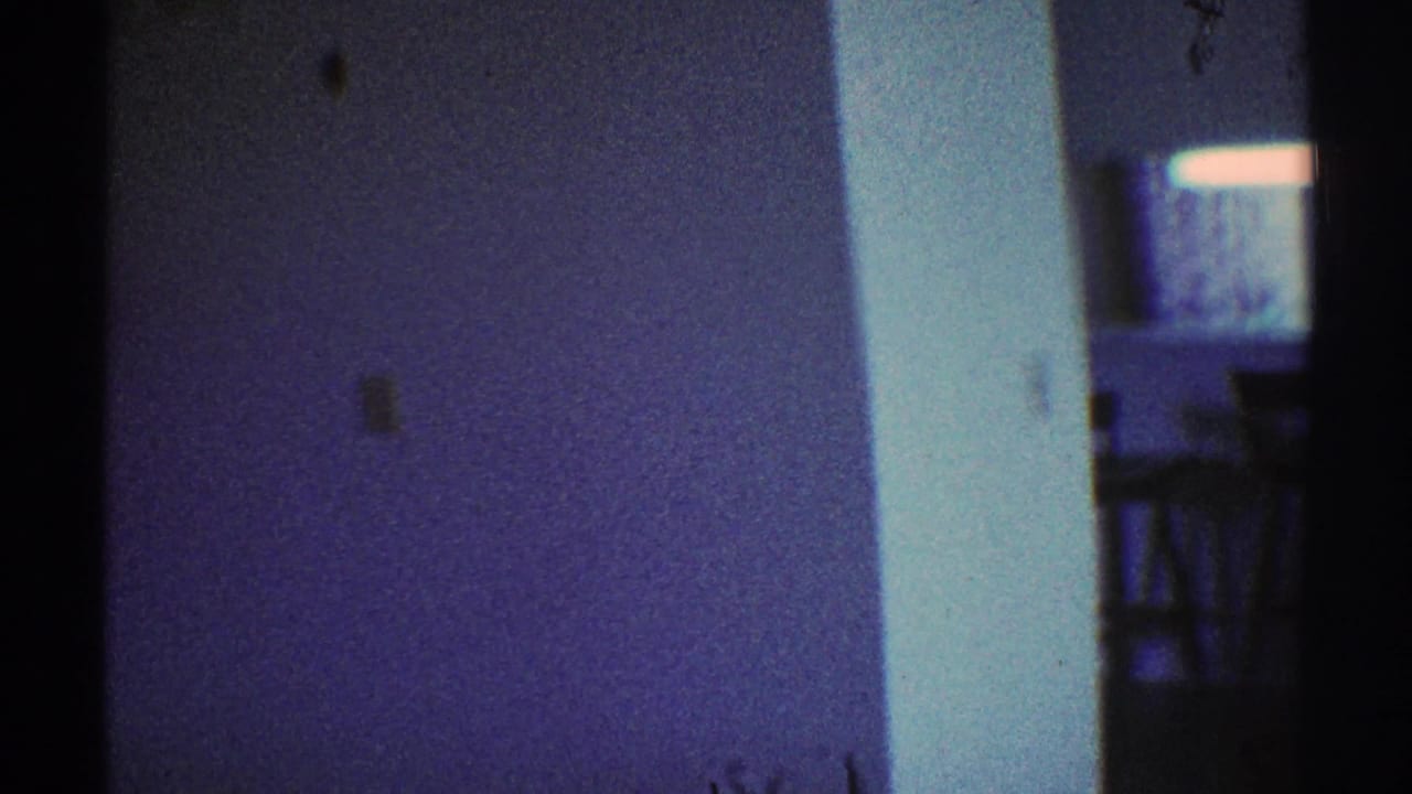 1969 Los Angeles home-movie frame showing a dim private interior with white walls, door trim, and a dark passageway.