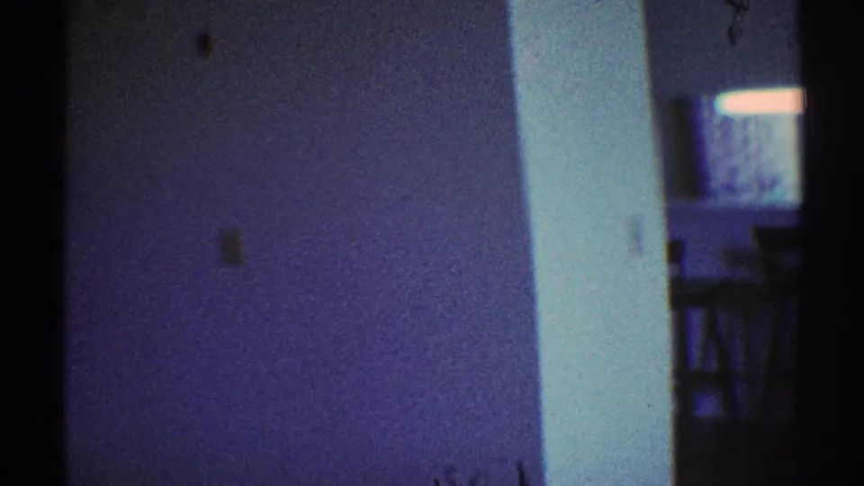 1969 Los Angeles home-movie frame showing a dim private interior with white walls, door trim, and a dark passageway.