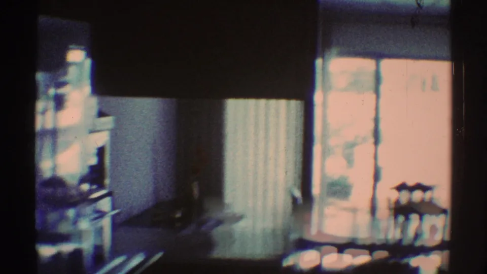 1969 Los Angeles home-movie frame showing a curtained glass door, small dining table, chairs, and kitchen area.