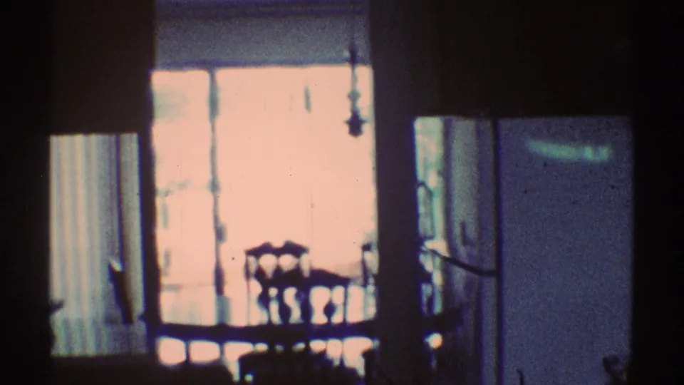 1969 Los Angeles 8mm frame of a dim dining area with wooden chairs and a table near a bright window, partly obscured by dark film damage.