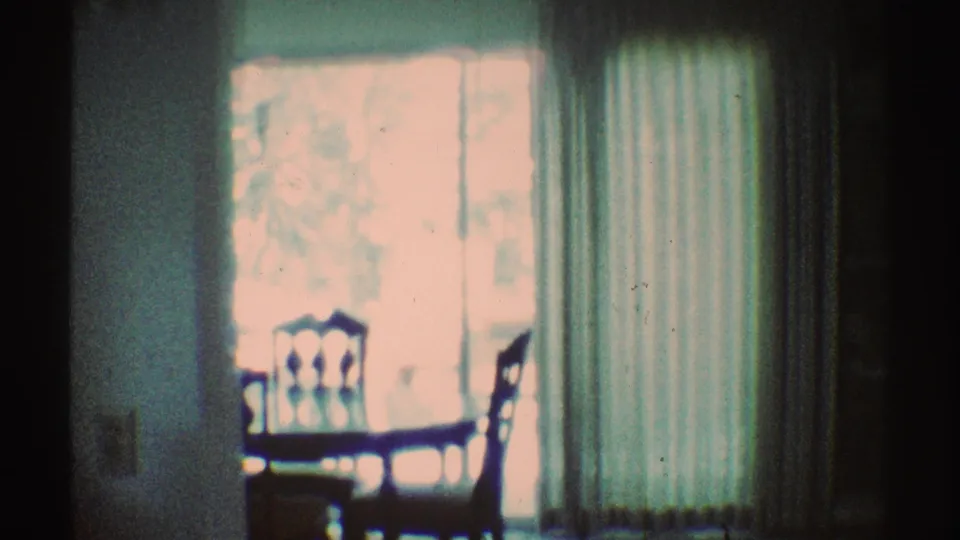 1969 Los Angeles 8mm frame showing a dining chair and table by a bright window, with vertical curtains and dark underexposed areas.