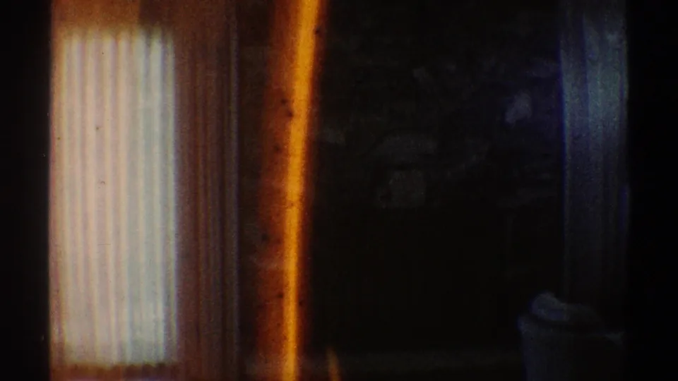 Damaged 1969 8mm interior frame with vertical curtains, deep shadows, and a warm burn-like streak interrupting the view.