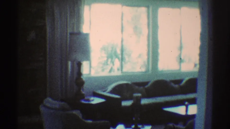 1969 Los Angeles 8mm frame of a living room with a sofa, lamp, curtains, and bright windows, affected by color shift and dark film bands.