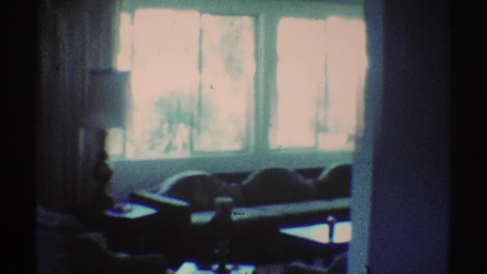 1969 Los Angeles home-movie frame showing a dim living room with bright windows, sofa shapes, a table, and a lamp.