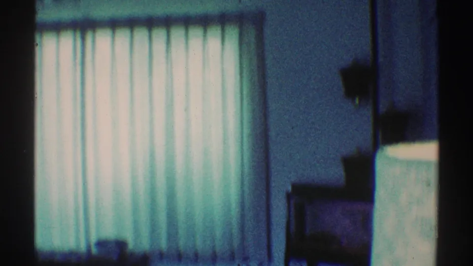 Blue-tinted 8mm frame of a private room corner with vertical curtains, wall space, and shadowed furniture along the right side.