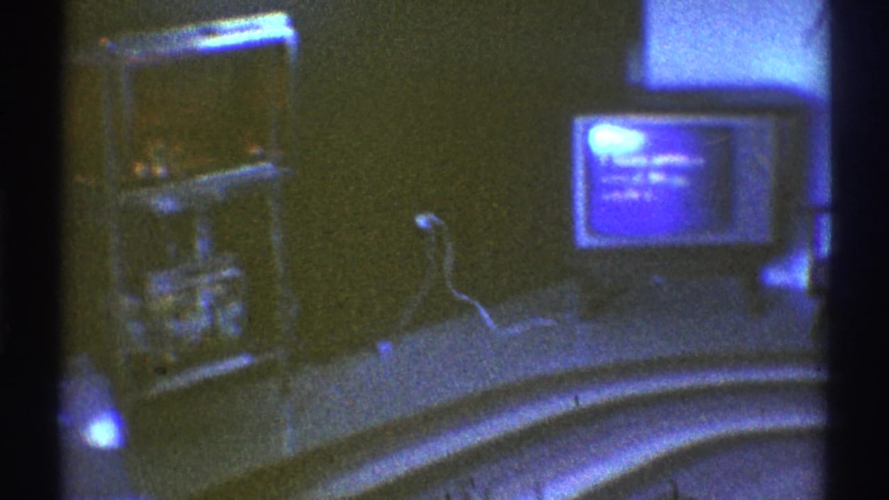 1969 Los Angeles home-movie frame showing a dim living room with a box television, shelving, curved table, and cool blue cast.