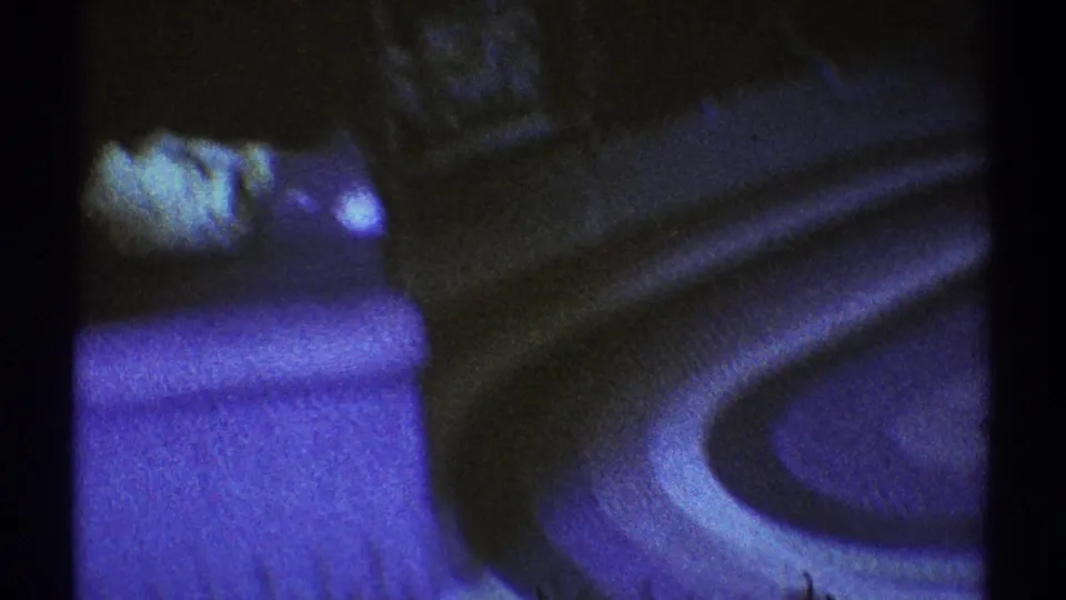 1969 Los Angeles living-room frame showing an upholstered chair, curved table, carpet, and dark furniture in low blue light.