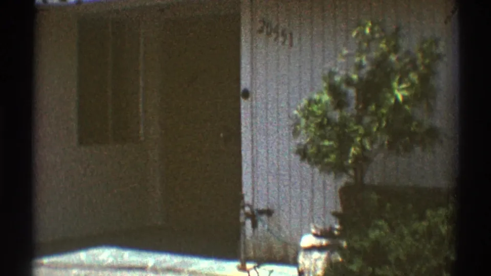 Los Angeles house exterior from 1969 home-movie footage, showing a front door, porch wall, potted shrub, and sunlit entry area.