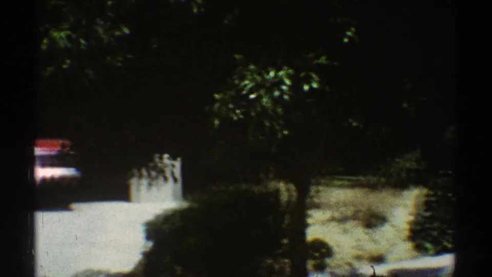 1969 Los Angeles residential frame with a red car glimpsed near a driveway, white wall, trees, and hedges.