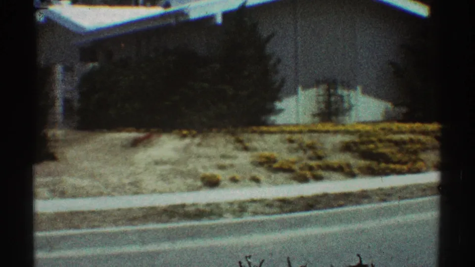 1969 Los Angeles home-movie frame showing a white residential exterior, front yard, curb, and low plantings.