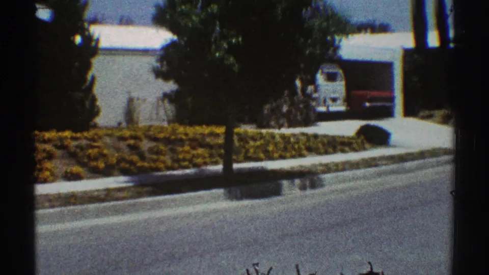 1969 Los Angeles home-movie frame showing a quiet suburban street, yellow flower bed, driveway, trees, and a white van beneath a carport.