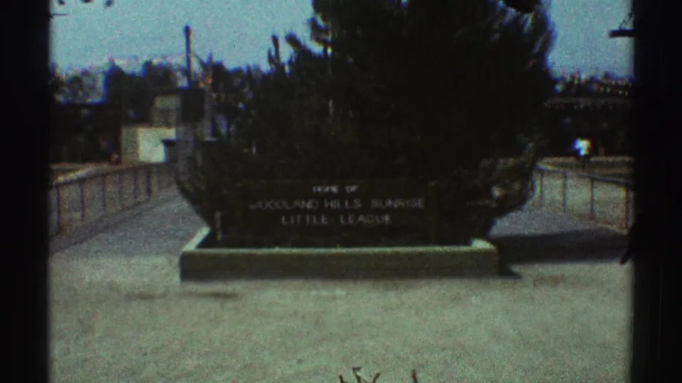 1969 Los Angeles frame showing a Woodland Hills Sunrise Little League sign near a sidewalk, landscaped planting, trees, and street railings.