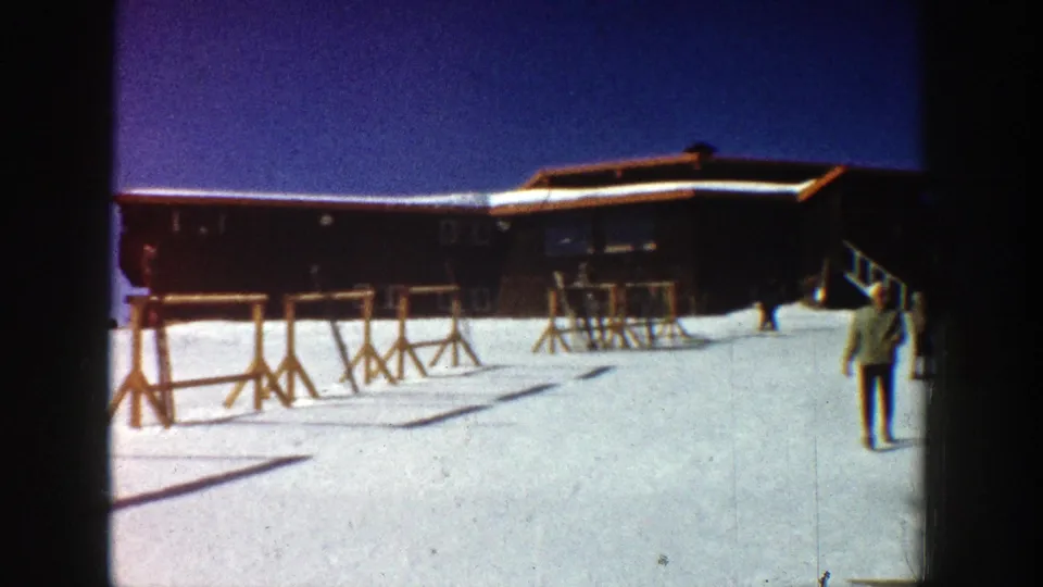 1961 Aspen ski resort scene with a wooden lodge, snow-covered ground, ski racks, and a person walking in winter clothing.