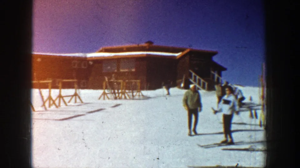 1961 Aspen resort view with two people in winter clothing near a lodge and ski racks on bright snow.