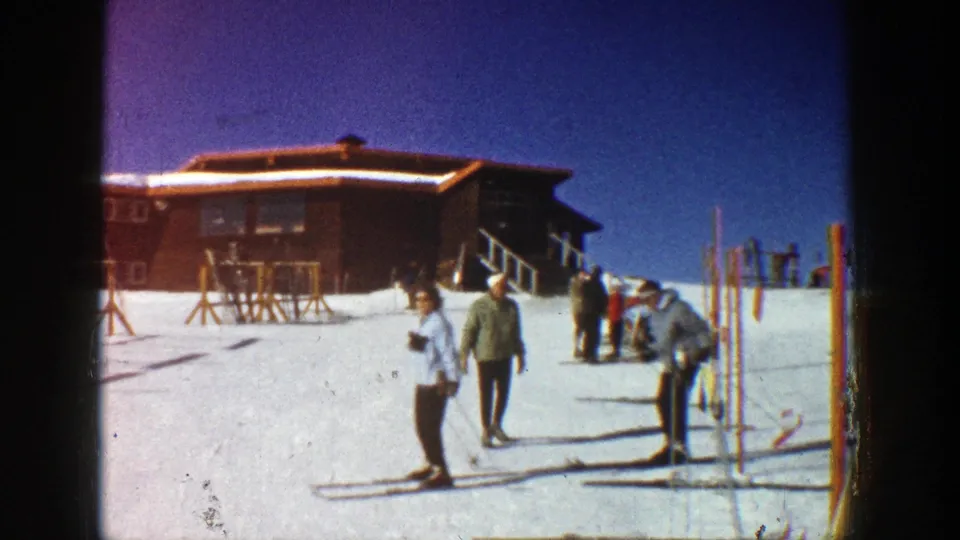 1961 Aspen ski area with visitors near upright poles, a wooden lodge, snow, and winter equipment.
