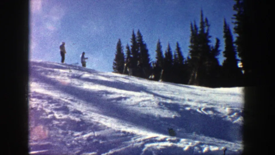 1961 Aspen winter home-movie frame showing two distant people on a sunlit snowy slope near evergreen trees.