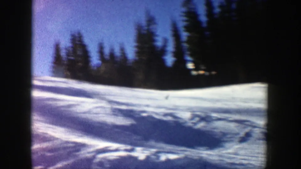 Soft 8mm frame of a bright Aspen ski slope in 1961, showing snow, tracks, and a dark evergreen background.