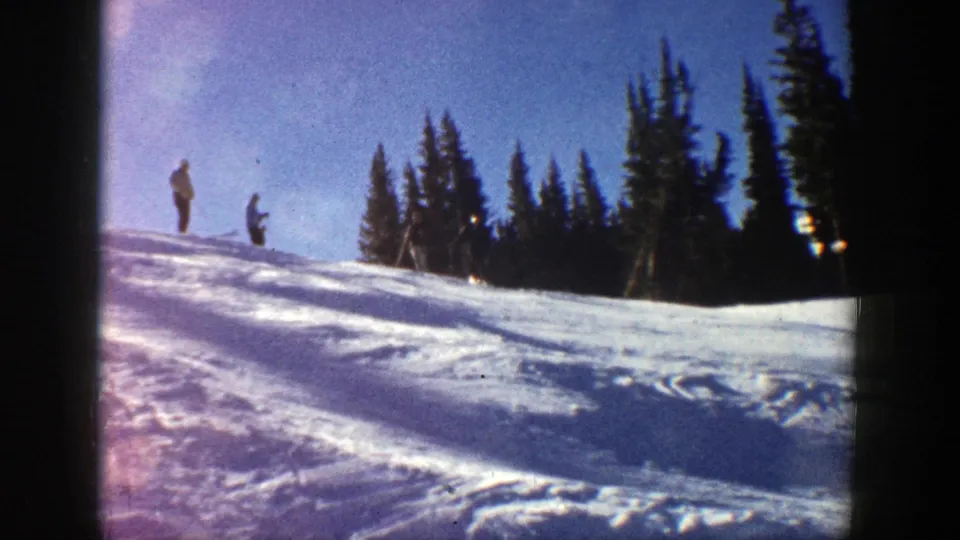 1961 Aspen frame with two distant skiers on a sunny snow-covered ridge, framed by evergreen trees and blue sky.