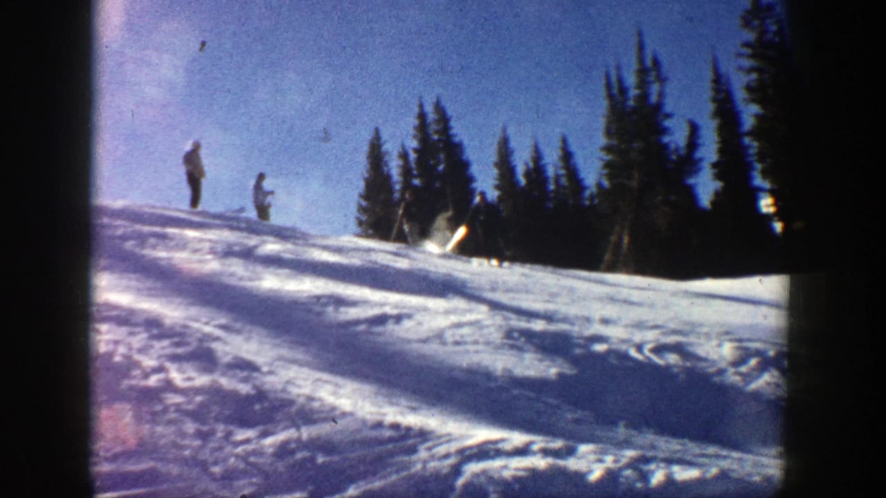 1961 Aspen home-movie frame showing two distant skiers on an icy snow slope with evergreens and blue sky behind them.