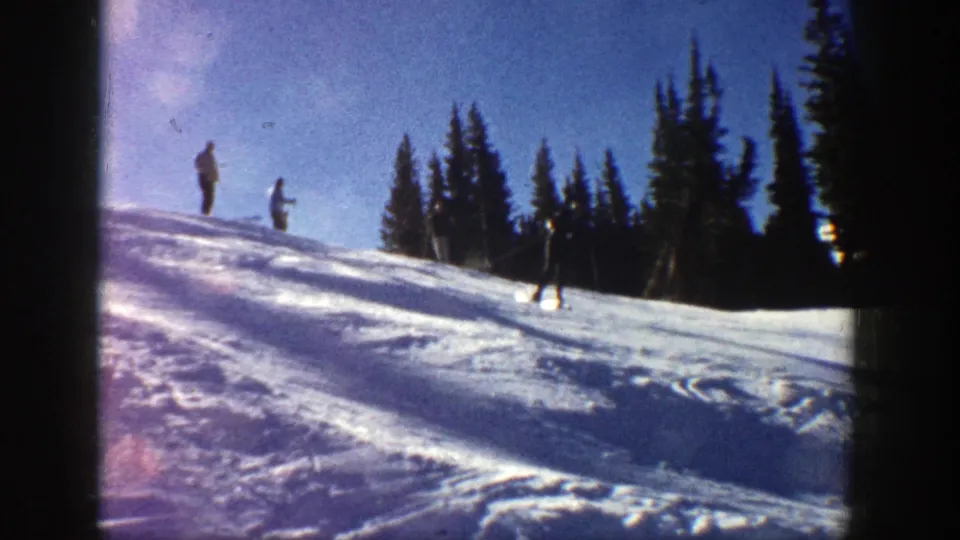 1961 Aspen ski footage with one skier descending across the snow while two figures remain near the top of the slope.