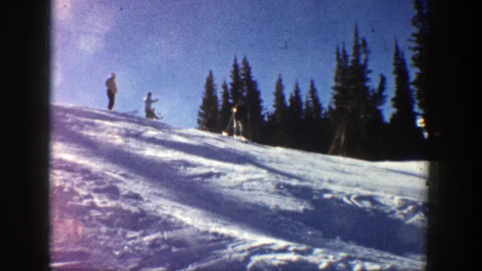 1961 Aspen home-movie frame showing distant skiers standing on a sunlit snowy slope with dark evergreens behind them.
