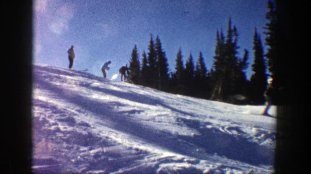 In 1961 Aspen home-movie footage, several men appear on a sunlit snowy ski slope near dark evergreen trees.