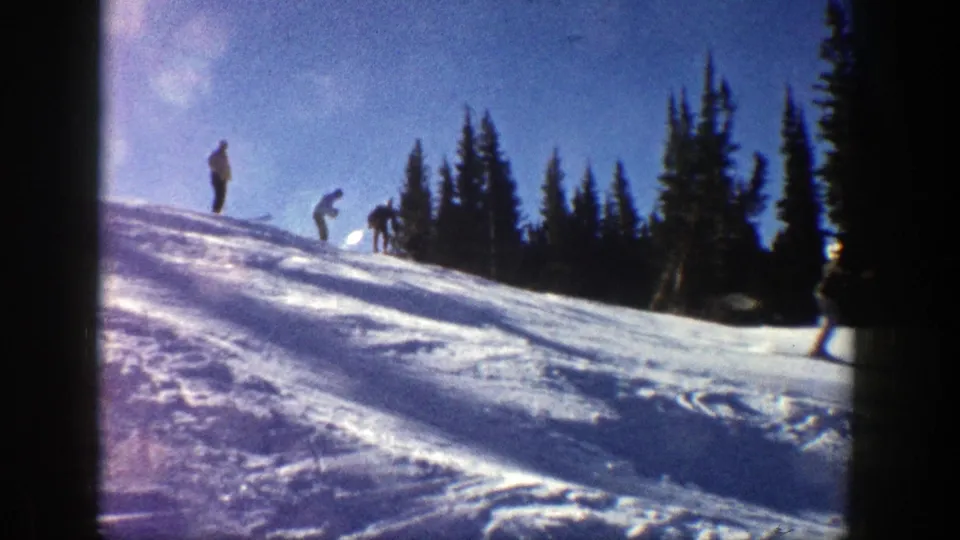 In 1961 Aspen home-movie footage, several men appear on a sunlit snowy ski slope near dark evergreen trees.