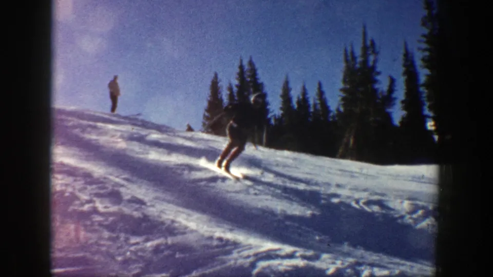 A skier moves downhill across a sunlit Aspen slope in 1961 home-movie footage, with trees silhouetted behind.