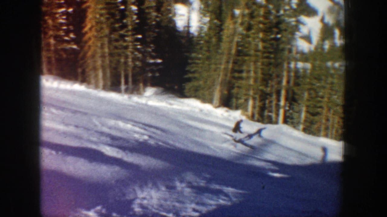 Skier descending near the right side of a snowy slope lined with evergreens.