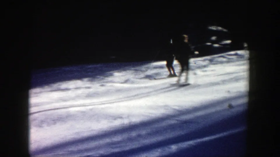 Distant skiers on a 1961 Aspen snowy slope, partly silhouetted against bright snow and forest shadows in an 8mm home-movie frame.