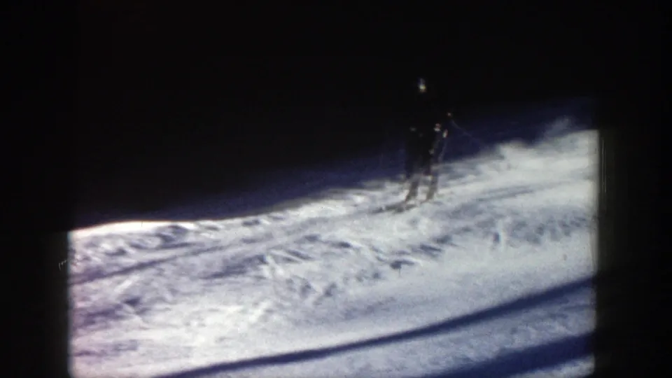 A skier descends a snowy Aspen slope in 1961, framed by dark evergreen shadows and bright tracked snow in 8mm footage.