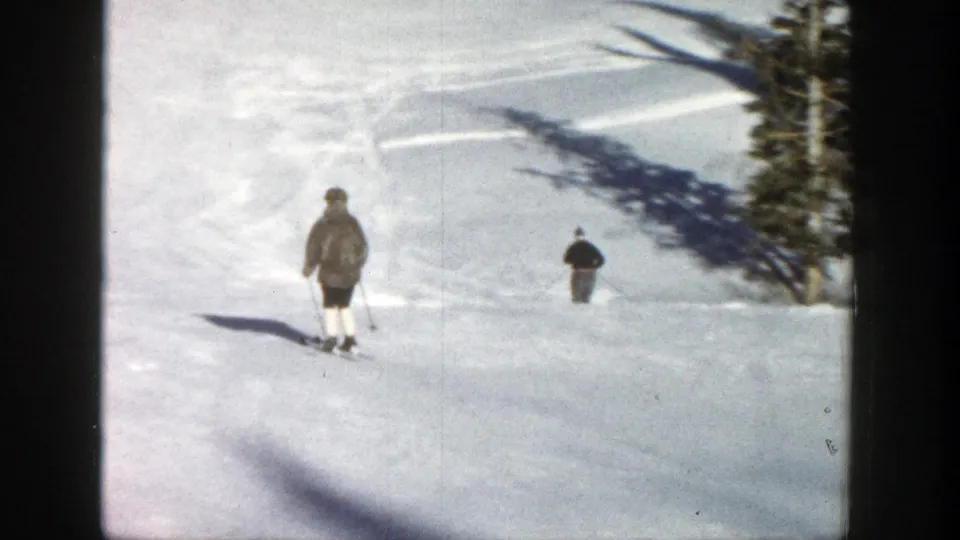 1961 Aspen frame with two distant skiers moving down a broad snow-covered slope, bordered by evergreens and ski tracks.