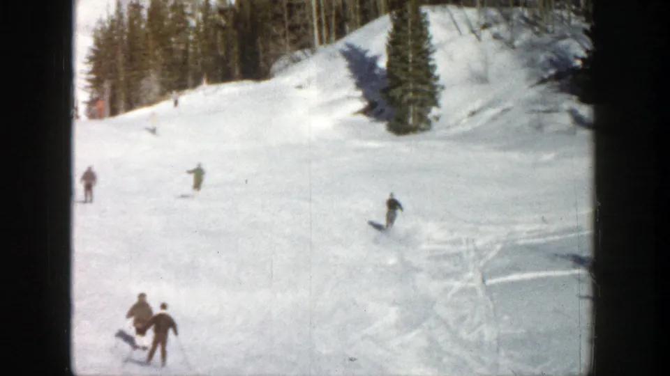 Wide 1961 Aspen frame showing several skiers on a snow-covered run, evergreen trees, bright snow, and visible 8mm contact-frame edges.