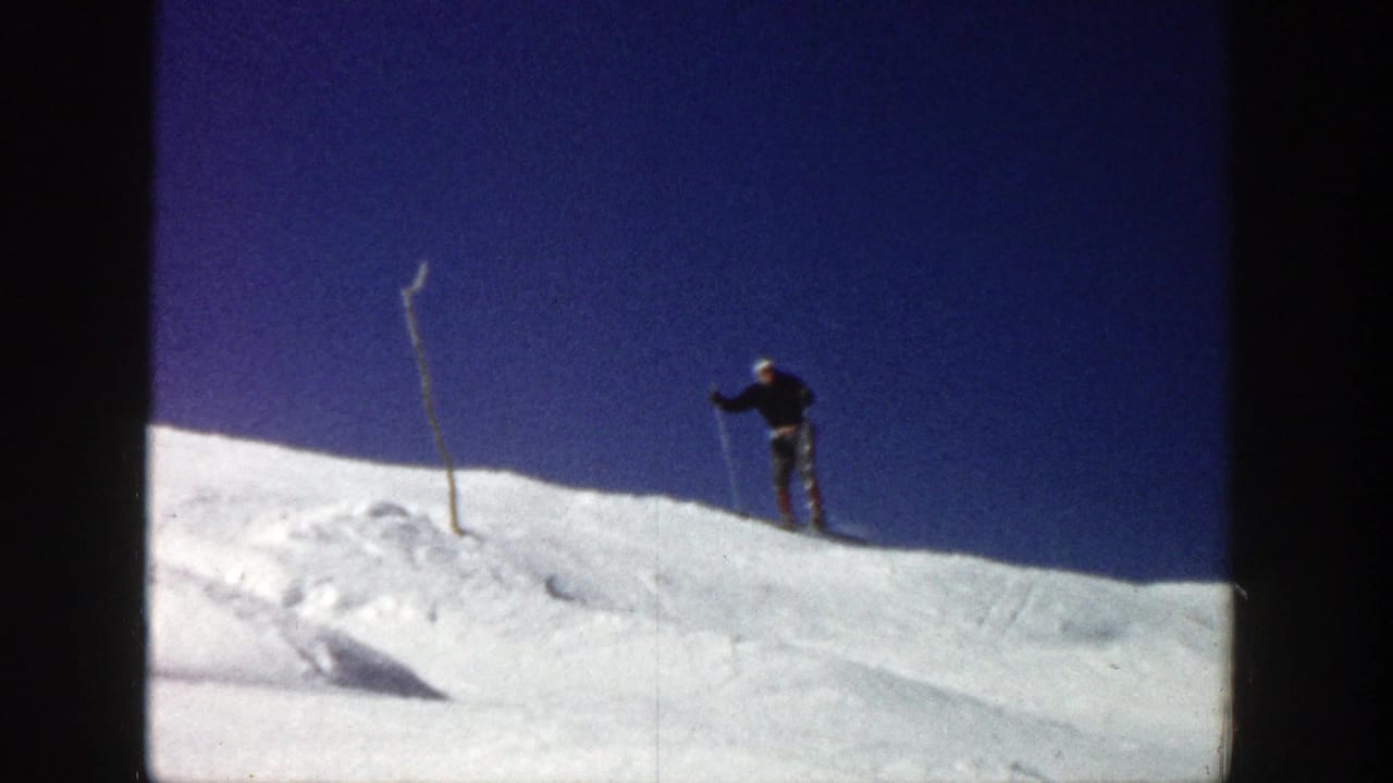 In 1961 Aspen home-movie footage, a skier in a dark jacket and light hat stands at the top of a snowy slope with poles nearby.