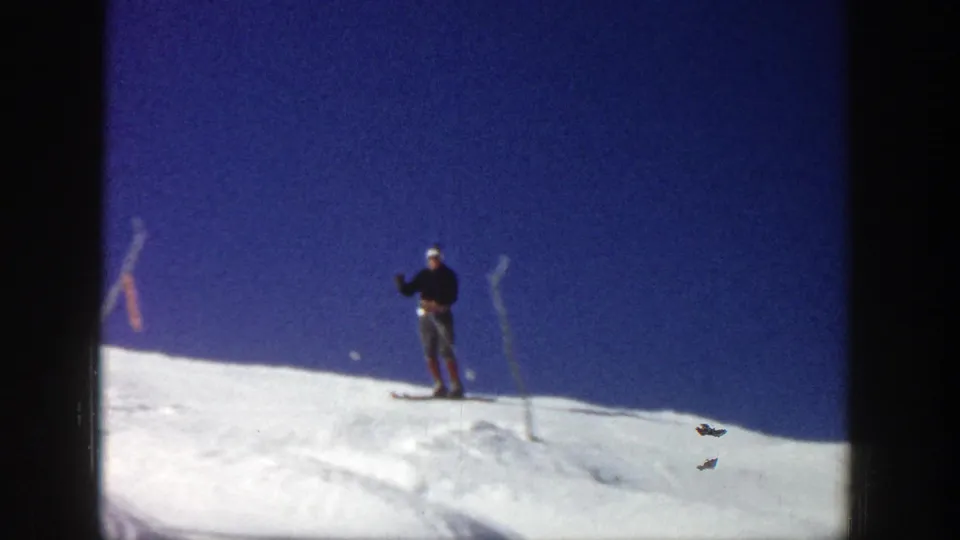 The Aspen skier stands near the hilltop on skis, framed by bright snow, a dark blue sky, and simple slope markers.