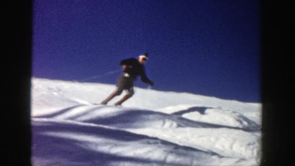 A skier in dark outerwear leans into a downhill turn on the Aspen slope, with textured snow and long shadows visible in the frame.