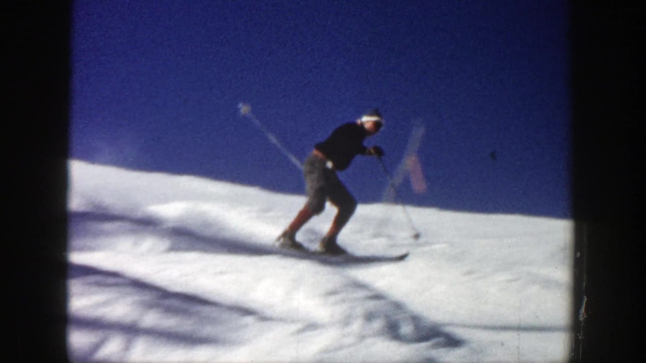 In 1961 Aspen footage, a man in a dark sweater and cap skis across sunlit snow with poles extended.