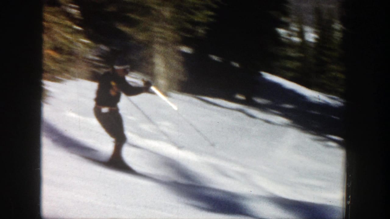 Skier in dark winter clothing moves across a snowy slope beside fir trees.