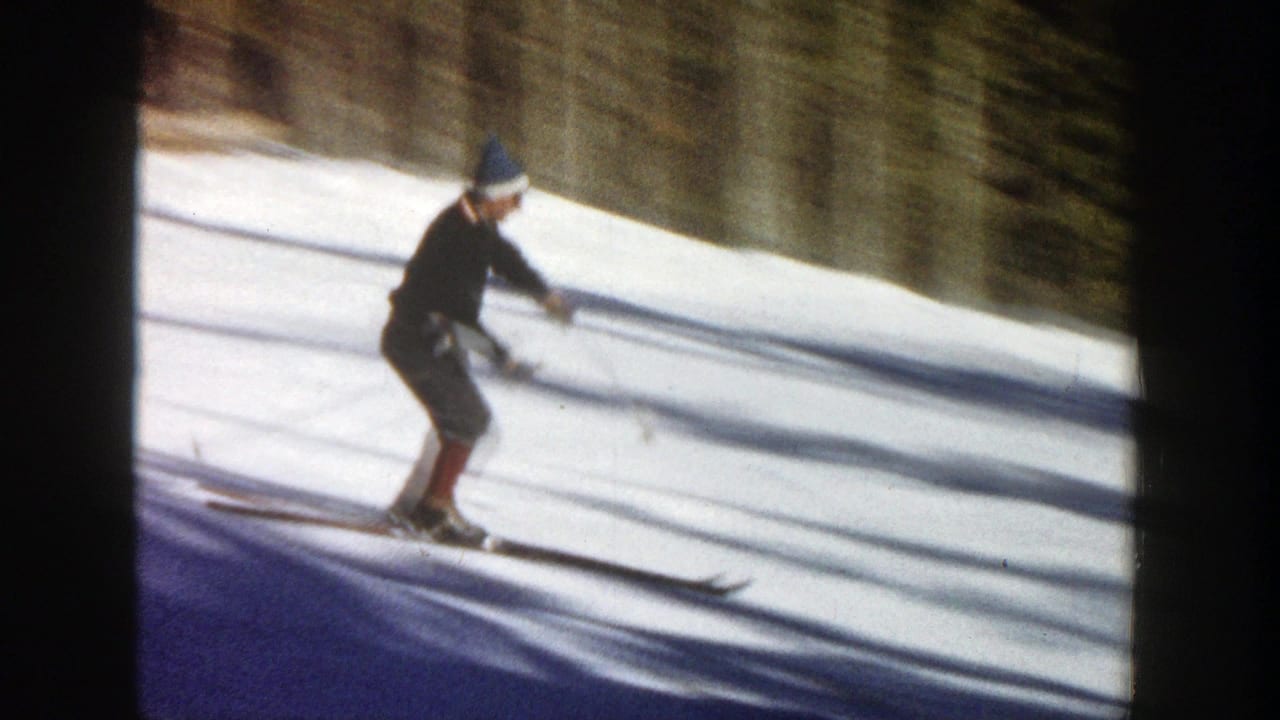 Skier in a hat and dark jacket travels across bright snow with long shadows.