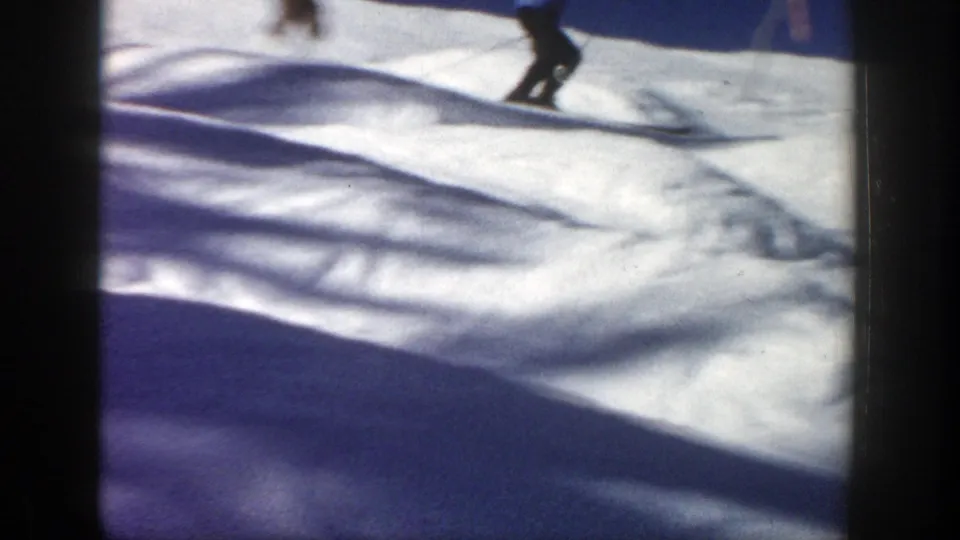 A motion-blurred skier moves down uneven snow in 1961 Aspen footage, partly obscured by shadow and frame softness.