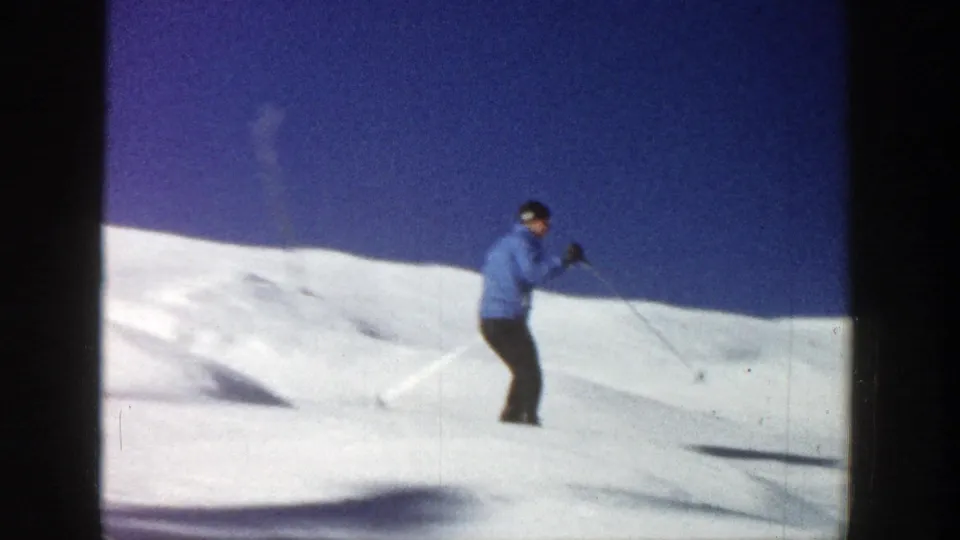 A skier in a blue jacket crosses an open snowy slope in Aspen, Colorado, with a deep blue sky above the snowbank.