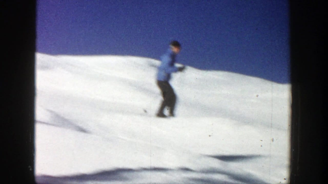 Skier in a blue jacket crosses a bright snowy slope under a clear sky.