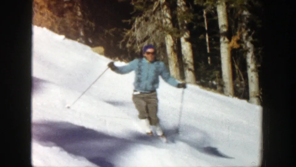 Adult skier in a light blue jacket faces the camera while descending a snowy Aspen slope bordered by evergreens.