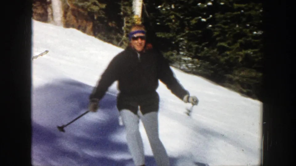 Adult skier in a dark jacket and light pants passes near the camera on a wooded snowy slope in 1961 Aspen footage.