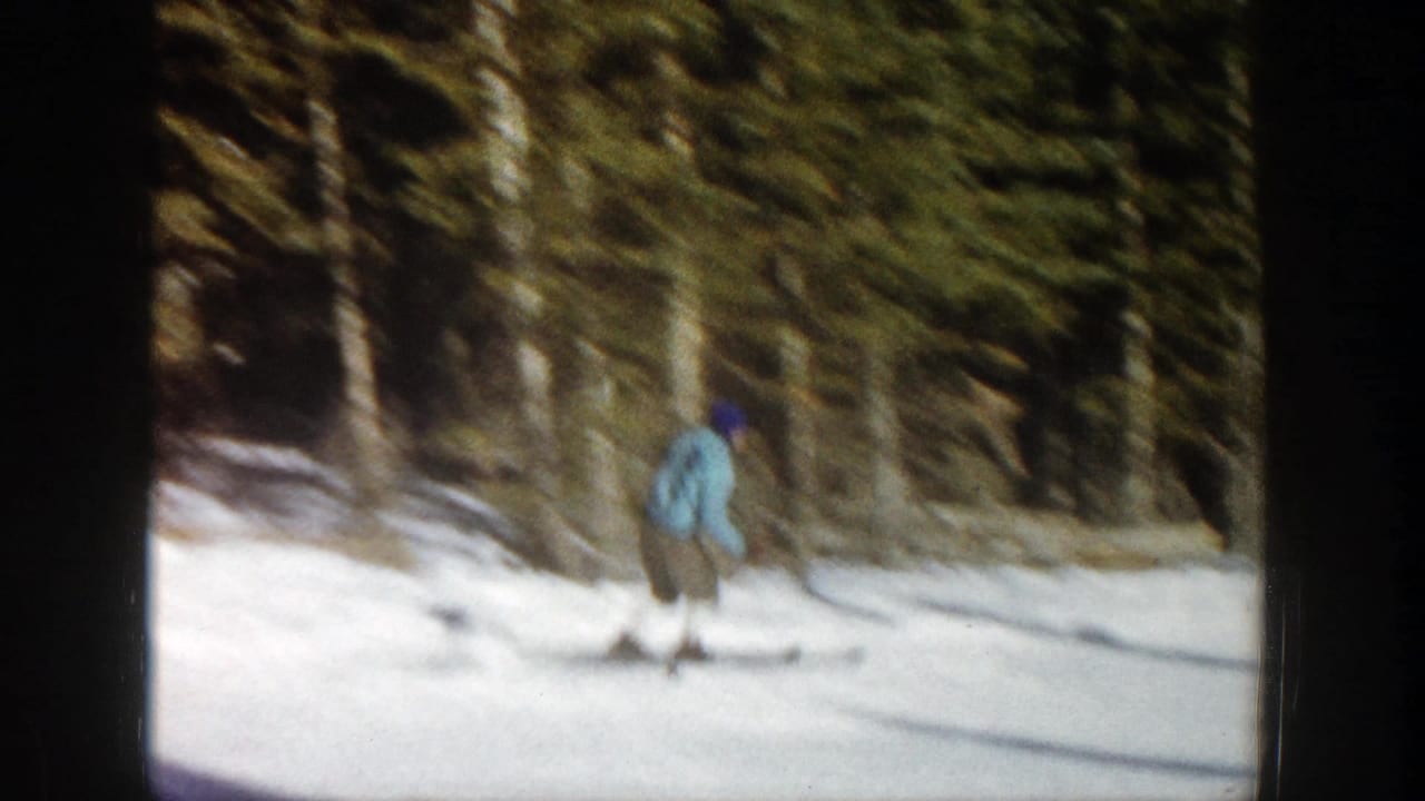 A skier in a light blue jacket moves across a snowy slope bordered by evergreen trees.