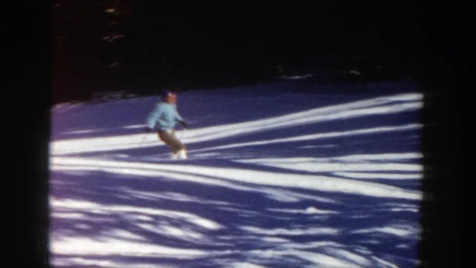 1961 Aspen home-movie frame with a skier leaning into motion on a snowy slope crossed by tree shadows.