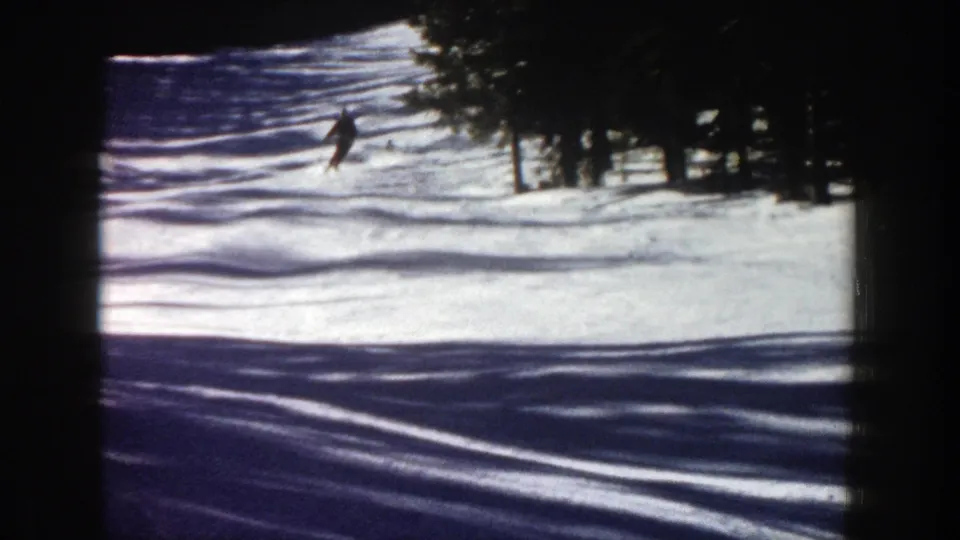 1961 Aspen frame showing a distant skier descending a snow-covered run framed by evergreen trees and long shadows.